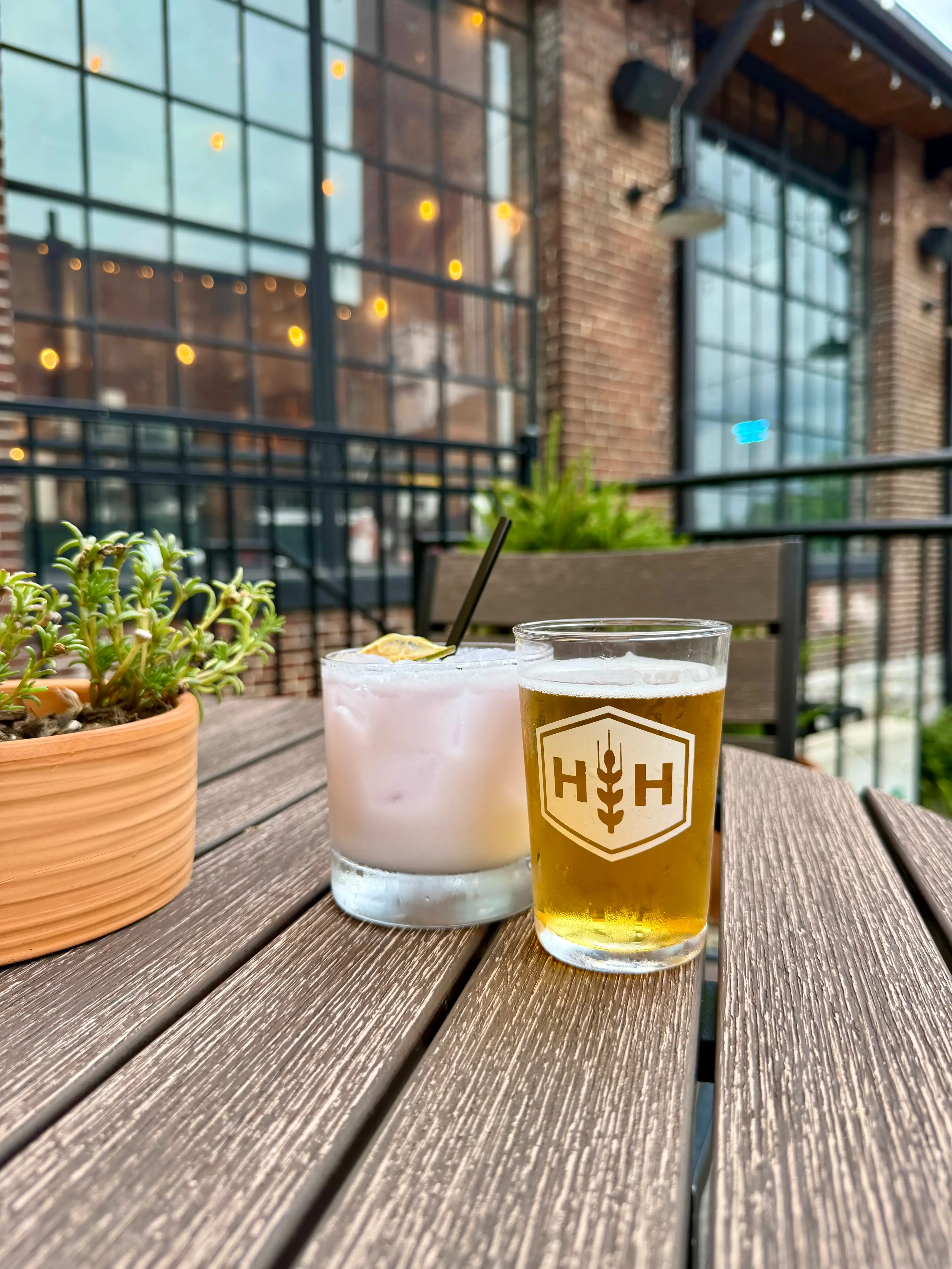 A pale cocktail with a dried citrus garnish next to a Harding House beer on a patio table, with string lights and factory windows behind them.