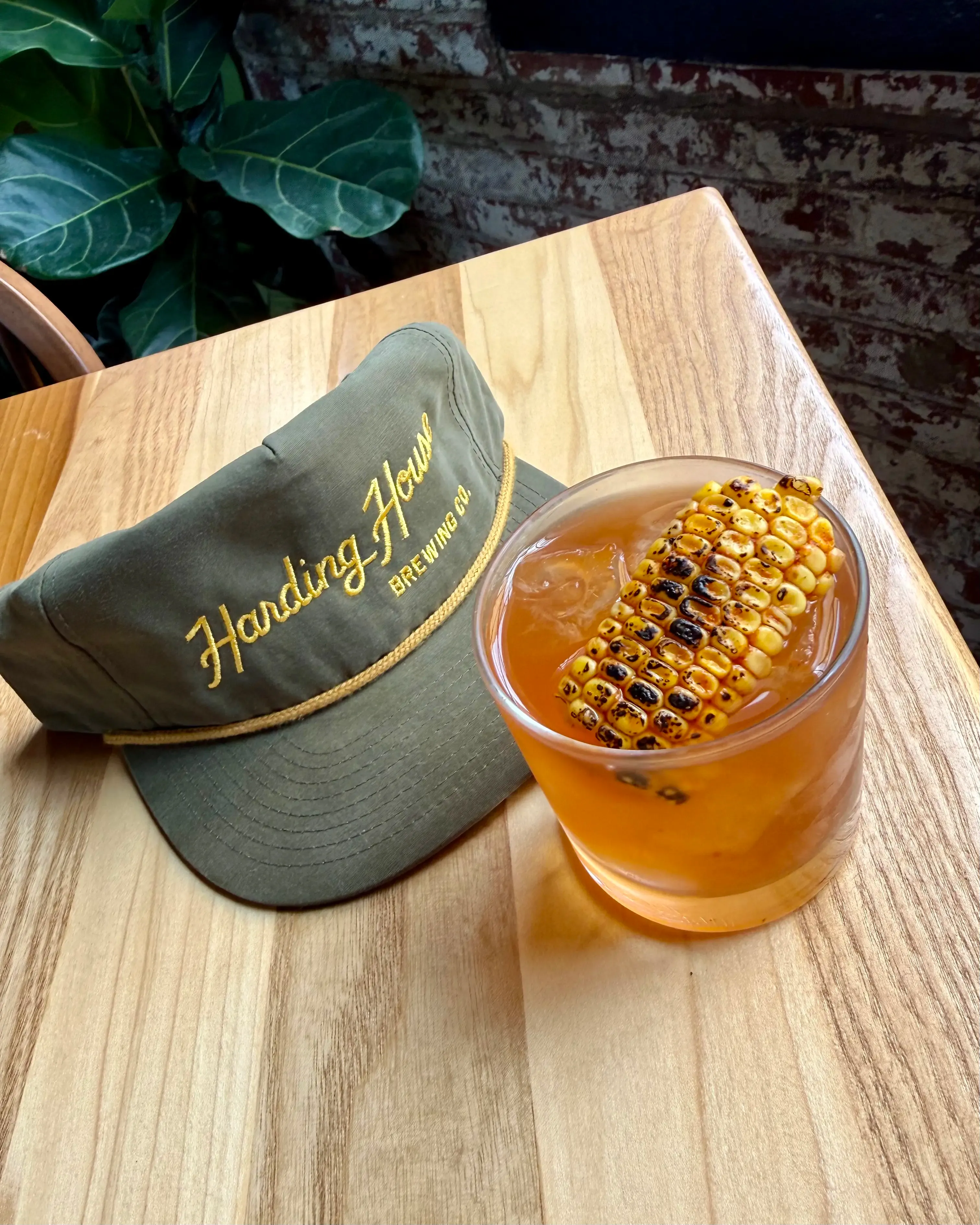 A cocktail garnished with charred sweet corn next to a Harding House hat on a wooden table.
