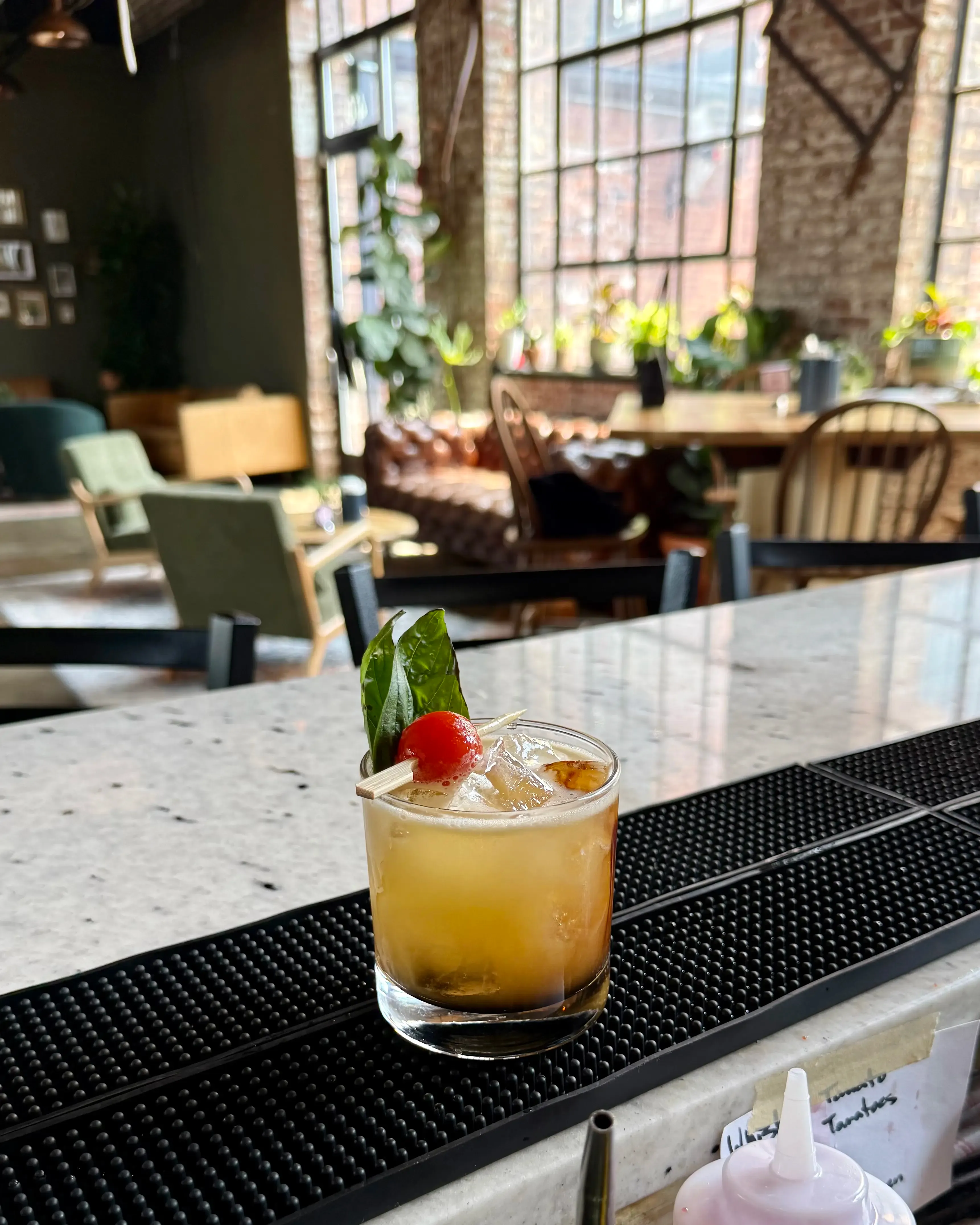 A cocktail garnished with basil and a cherry tomato on the bar, with the lounge and tall factory windows behind it.