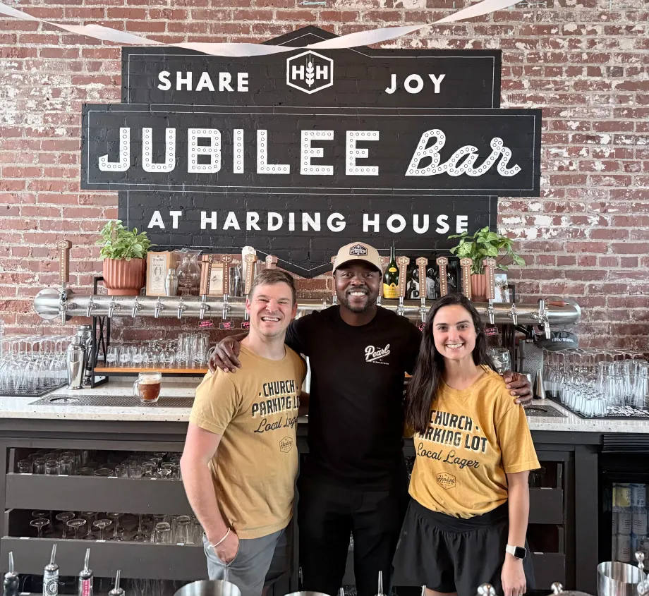 Nate, Taylor, and Akinde behind the bar at Harding House, in front of the painted Jubilee Bar sign.