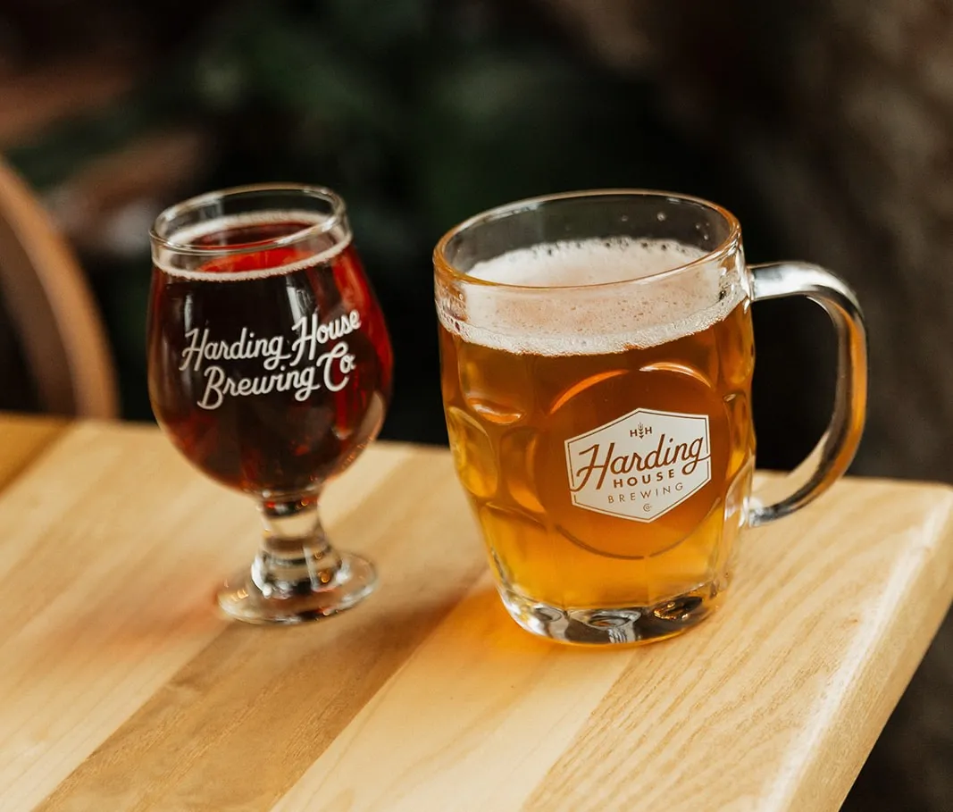 A dark beer in a Harding House Brewing Co. goblet next to a golden beer in a dimpled Harding House mug on a wooden table.